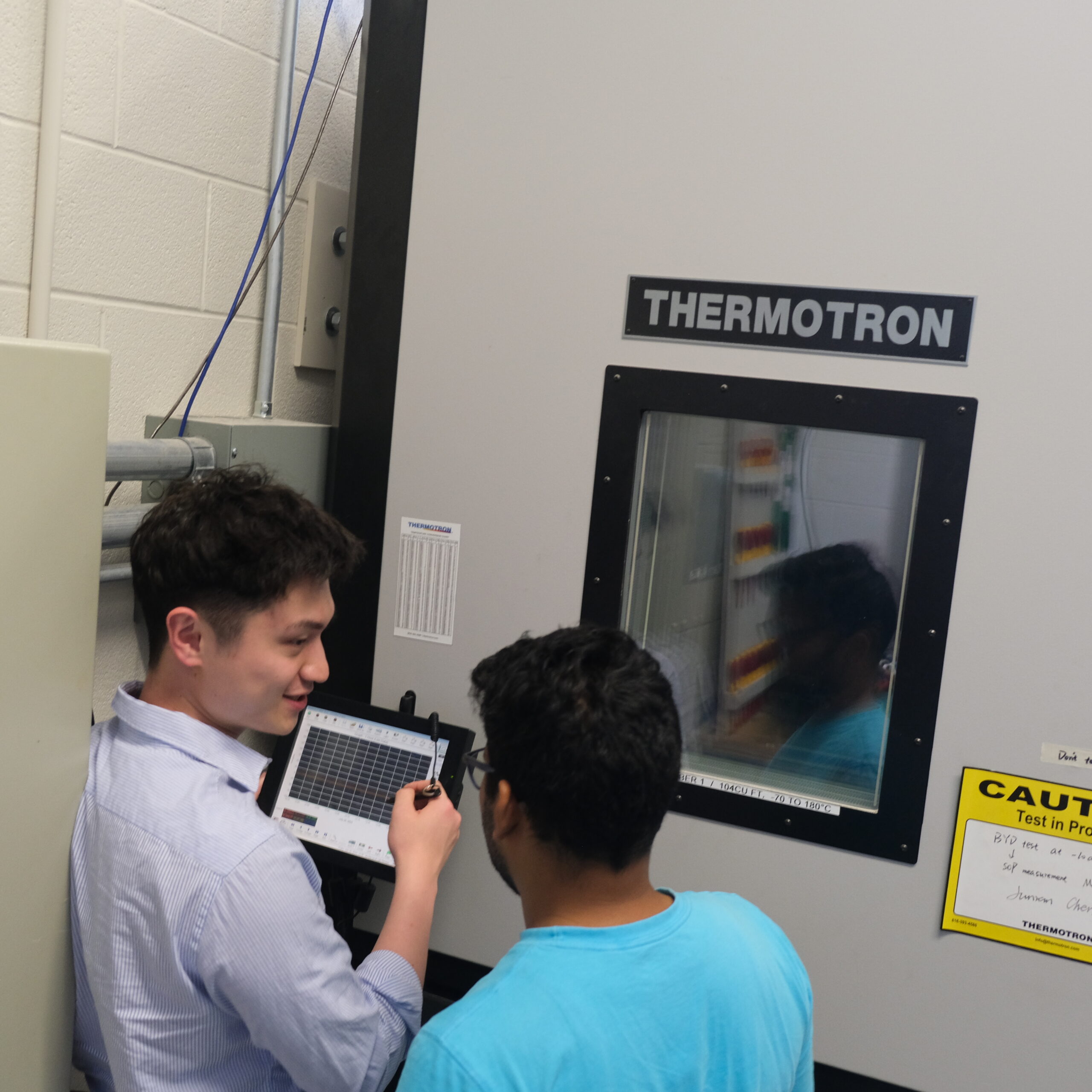 Junran (left) and Biswanath (right) in front of the large thermal chamber control panel. Junran is pointing out some of the functions of the device.