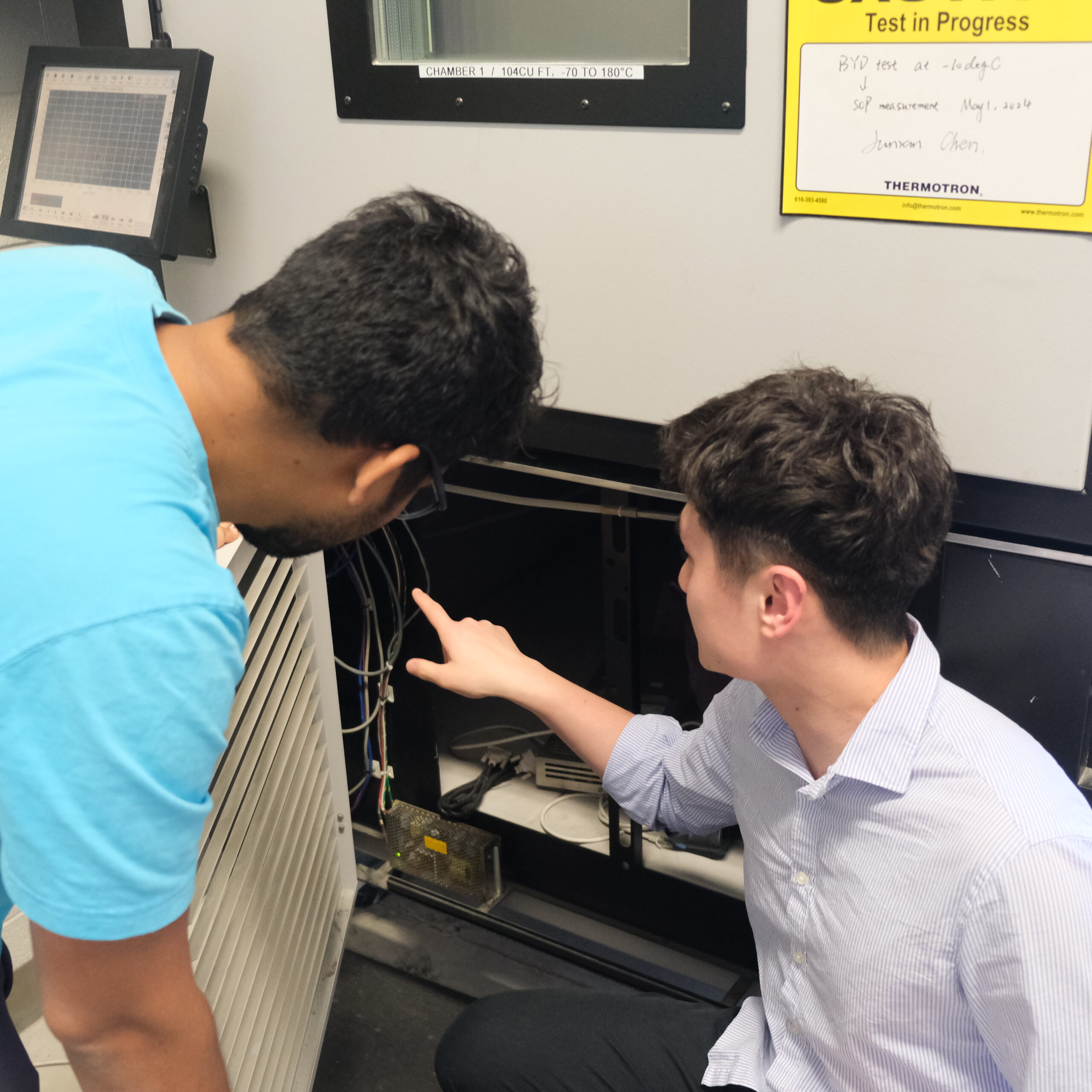 Biswanth (left) and Junran (Right) looking into the underbelly of the large chamber, where the microcontroller is located. There is a yellow sign in front of the equipment stating 'test in progress'.