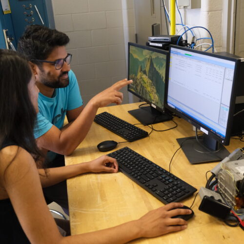 Two research students, Sobhika on the left and Biswanath on the right, working at a desk with two monitors. Biswanath is pointing at the Arbin control panel monitor as they make adjustments to the battery cycler schedule