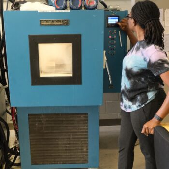 Lucia standing beside a small blue chamber, adjusting the temperature settings on the control panel. The image provides a size comparison of the equipment.