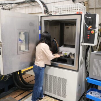 Qi looking inside a large chamber as she sets it up for testing. The chamber door is open, and she is arranging equipment inside.