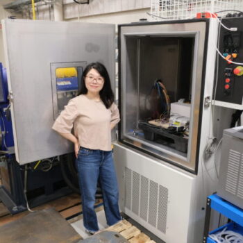 Qi standing in front of a large 20 cubic foot chamber with its door open, highlighting its size. Inside the chamber, a module is being set up for testing.