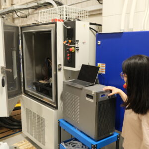 Qi turning on a PolyScience chiller which is located beside the 24 square foot module test chamber in the MARC 117 lab. There is a laptop resting on top of the chiller and the chamber door is open.